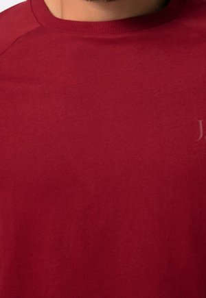 Red cotton T-shirt with a round neckline, visible stitching, and a subtle logo detail on the chest. Smooth texture, casual design.