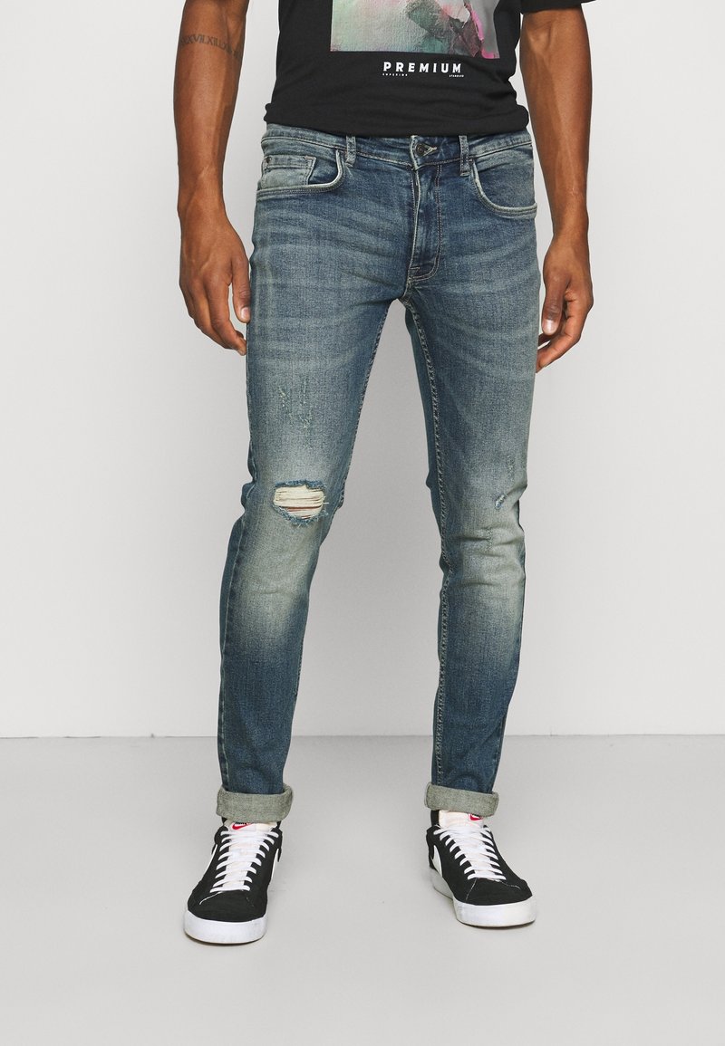 Homme portant un jean bleu délavé avec des bas retroussés, un genou déchiré sur la jambe gauche, des baskets noires avec des lacets blancs et un t-shirt noir.