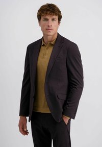 Mann mit lockigem braunem Haar, der ein dunkles Blazer, ein senfgelbes Strickpolo-Shirt und dunkle Hosen trägt, steht mit einer Hand in der Tasche.