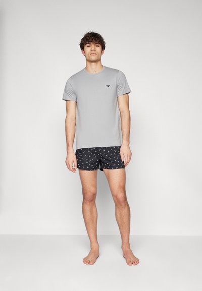 Grau Kurzarm-T-Shirt mit schwarzem Logo, kombiniert mit schwarzen Badeshorts, die ein kleines weißes geometrisches Muster aufweisen. Modell barfuß und stehend.