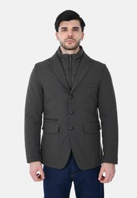 Giacca grigio scuro con una texture liscia, caratterizzata da una chiusura frontale con bottoni, due grandi tasche frontali e uno strato interno staccabile con zip.