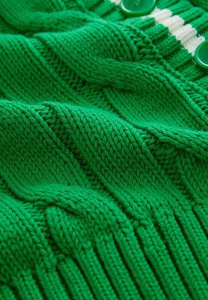 Zaļš adīts audums ar teksturētām rakstām, kurā ir ribotas daļas un gludas zonas, papildināts ar zaļām pogām un krēmkrāsas svītrām.