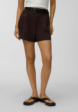 Femme portant un short plissé marron foncé taille haute avec une ceinture noire, un haut blanc et des sandales plateformes noires debout sur un sol gris clair.