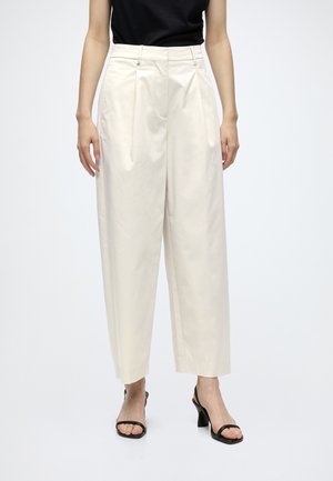 Femme portant un pantalon crème à jambes larges, un haut noir sans manches et des sandales noires ouvertes à talons, debout devant un fond uni.