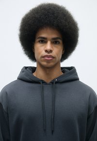 Siva kapuca s dvostrukim vezanjem i bočnimi šavovi, ki se ponaša z mehko teksturo. Model ima poln, kodrast afro frizuro pred enostavno ozadje.
