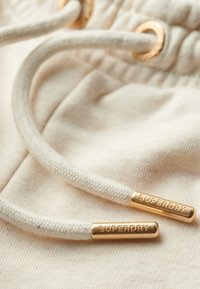 Cordon de serrage crème avec des embouts en métal doré marqués « SUPERDRY », attachés à un tissu doux et texturé de couleur claire.