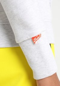 Cappotto grigio con manica a costine, con un'etichetta rossa "Superdry". Tessuto giallo brillante visibile sotto. Materiale morbido.