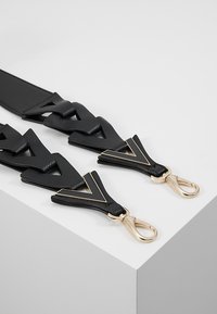 Svart läderrem med V-formade utskärningar, prydd med hardware i guldig färg och kräftklor. Texturerad yta ger kontrast.