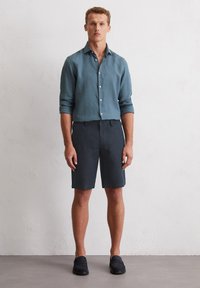 Men's lightweight, unbuttoned, teal linen shirt, paired with dark shorts. The outfit features a relaxed fit and texture with simple hardware.
