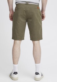 Olivgrüne Shorts mit glatter Textur, zwei Gesäßtaschen und einem Lederetikett. Kombiniert mit weißen Socken und Sneakers mit einem schwarzen Logo.