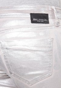 Jean en métal argenté avec une poche arrière portant une étiquette noire indiquant "Mavi Jeans Co." Des coutures blanches soulignent la poche.