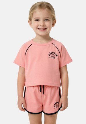 Jeune fille souriante portant un t-shirt et un short roses assortis de la marque ellesse club avec une bordure noire, debout sur un fond blanc.