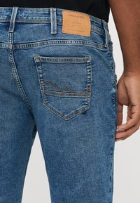 Kadar izbliza osobe koja nosi plave traperice s kožnom oznakom marke Jack & Jones na pojasu i vidljivim šavovima stražnjeg džepa.