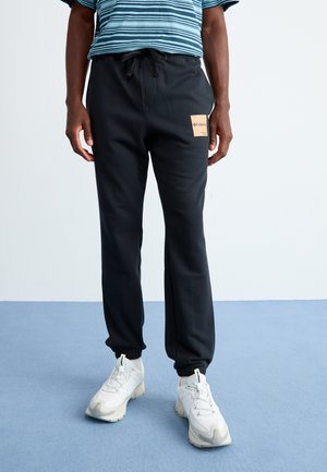 Person iført sorte sweatpants med et beige Columbia-logo patch, hvide sneakers og en blåstribet skjorte, der står på et blåt gulv.