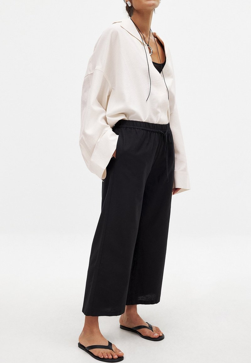 Oversized crèmekleurige blouse met een kraag, zwarte broek met elastische tailleband en zwarte flip-flops, tegen een effen witte achtergrond.