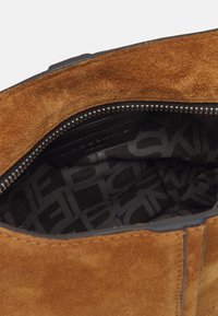 Ruskea mokkahuopakassi, jossa on vetoketjukiinnitys, musta kankaallinen vuori, jossa on toistuva logokuviot. Näkyvissä on teksturoitu pinta.