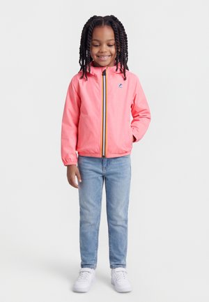 Hymyilevä lapsi, jolla on punotut hiukset ja päällään kirkkaan korallinvärinen vetoketjullinen takki, vaaleat farkut ja valkoiset tennarit, seisoo yhdellä kädellä taskussa.