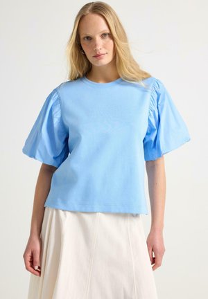 Donna che indossa un top azzurro con maniche a sbuffo e una gonna avorio, in piedi davanti a uno sfondo bianco semplice, che guarda verso la fotocamera.