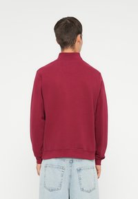 Pulover din burgund cu guler înalt, mansete și tiv ribat. Textură moale cu detalii minime, purtat cu pantaloni scurți din denim de culoare albastră deschisă.