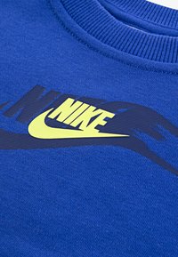 Mėlyna džemperis su raukšlėta apykakle. Pasižymi dideliu geltonu "Nike" logotipu ir juodais akcentais. Pagamintas iš tekstūruotos, minkštos medžiagos.