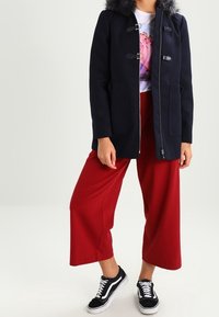 Marinblå ullblandad kappa med huva i konstpäls, utrustad med silverfärgade knäppningar och två framfickor, kombinerad med vida röda byxor och svarta sneakers.