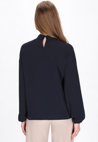 Blusa a maniche lunghe blu navy con apertura a chiave sul retro, vestibilità ampia e polsini arricciati. Tessuto morbido con texture minima.