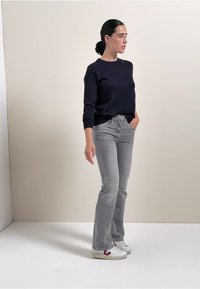 Marine Crewneck-Sweatshirt, hellgraue ausgestellte Jeans, weiße Sneaker mit dunklen Akzenten. Modell steht vor einem schlichten hellen Hintergrund.