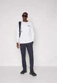 Ilgas baltas marškinėlis su juodu logotipu, tamsiai mėlynos kelnės cargo stiliaus, juodas kuprinė, tamsūs akiniai nuo saulės ir juodi sportbačiai. Paprasta fono spalva.