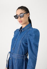 Vestido de ganga com cintura ajustada, mangas bufantes e frente com botões, combinado com óculos de sol angulares pretos e brincos de argola prateados.