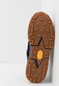 Semelle en caoutchouc avec un motif marron et noir, présentant un logo Vibram jaune et un crampon triangulaire pour une meilleure adhérence.