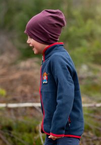 Marineblå fleecejakke med røde detaljer og et tegneseriepatch har en høj krave og lynlås. Barnet bærer en bordeaux farvet hue.