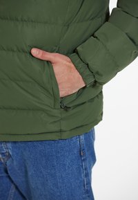 Grön puffjacka med strukturerad yta, utrustad med en dragkedjeförsedd sidoficka och elastiska ärmslut. Bärs över denimbyxor.
