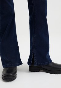 Tmavě modré rozšířené džíny s viditelným šitím a bočními rozparky na lemu, doplněné černými botami s gumovou podrážkou.