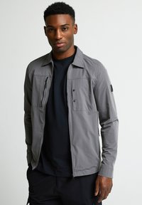 Pelēks vieglais jaka ar apkakli, divām priekšējām kabatām un rāvējslēdzēju. Valkāta uz melnas krekla, ar izsmalcinātu dizainu.