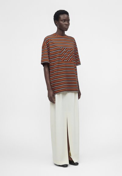 Γυναίκα που φοράει υπερμεγέθη ριγέ καφέ και μαύρη μπλούζα με τσέπη στο στήθος, συνδυασμένη με μακριά λευκή φούστα με σκίσιμο μπροστά και μαύρα παπούτσια.