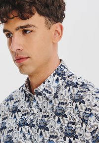 Biała koszula z zawiłym niebieskim i granatowym wzorem kwiatowym. Posiada standardowy kołnierz, zapięcie na guziki i lekki, przypominający bawełnę materiał.