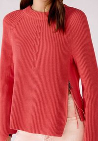 Pull en tricot rose corail avec une texture côtelée, un col rond et des zips sur les côtés pour un ajustement modulable. Le pull a une silhouette décontractée.