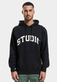 Svart hettegenser laget av bomull med en frontlomme, med stor hvit tekst "STUDIO" og en grønn detalj på brystet.