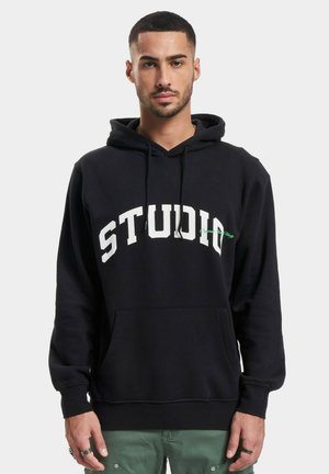 Svart hettegenser laget av bomull med en frontlomme, med stor hvit tekst "STUDIO" og en grønn detalj på brystet.