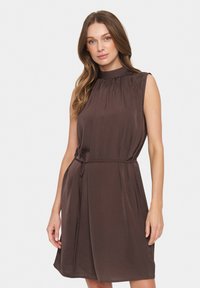 Rochie maro fără mâneci, cu guler înalt, textură moale și talie cu şnur. Prezintă pliseruri la decolteu și o siluetă fluidă.