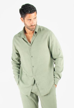 Chemise boutonnée vert clair en tissu doux et texturé, avec de longues manches et un col légèrement ouvert. Accent d'un bouton unique sur les manches.