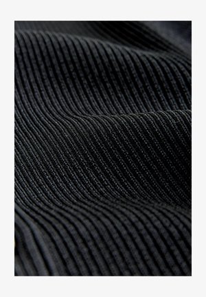 Lähikuva mustasta korduroy-kankaasta, jossa näkyy kohotettuja rinnakkain olevia uria ja pehmeä rakenne lempeine poimuineen.