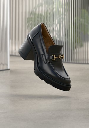 Mocassin à talon en cuir noir avec semelle épaisse et détail en métal doré, présenté sur un fond intérieur minimaliste avec des lignes verticales.