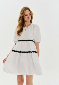 Biele šaty s pufovanými rukávmi a čiernym volánom, zvýrazneným pásom a rozšírenou sukňou. Hladká tkanina so štruktúrovaným siluetou.