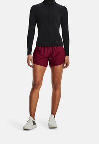 Schwarzes langärmeliges Oberteil mit Reißverschluss und hohem Kragen, kombiniert mit kastanienbraunen Sportshorts. Das Modell trägt weiße Turnschuhe mit grauen Akzenten.