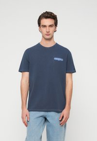 Μπλε βαμβακερό t-shirt με κοντά μανίκια, στρογγυλή λαιμόκοψη και μικρό μπλε γραφικό σχέδιο στο στήθος. Απαλή υφή, άνετη εφαρμογή.