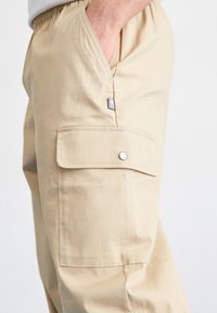 Pantalons cargo beiges avec taille élastique, poche latérale, poche à rabat avec fermeture à bouton-pression, tissu lisse et détails rebrodés.