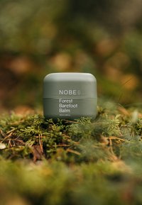 Grön cylindrisk behållare märkt "NOBE Forest Barefoot Balm", 100 ml, med en slät textur, placerad på mossig mark.