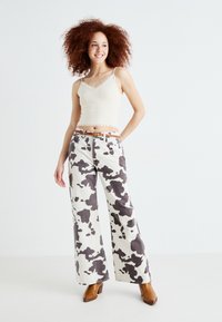 Jean large en imprimé vachette blanc et gris, associée à un débardeur léger beige avec bordure en dentelle. Des chaussures à talons marron complètent la tenue.