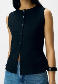 Ärmelloses schwarzes knappes Oberteil, das mit blauen Jeans getragen wird, mit einem schwarzen, klobigen Armband am rechten Handgelenk, vor einem schlichten hellen Hintergrund.